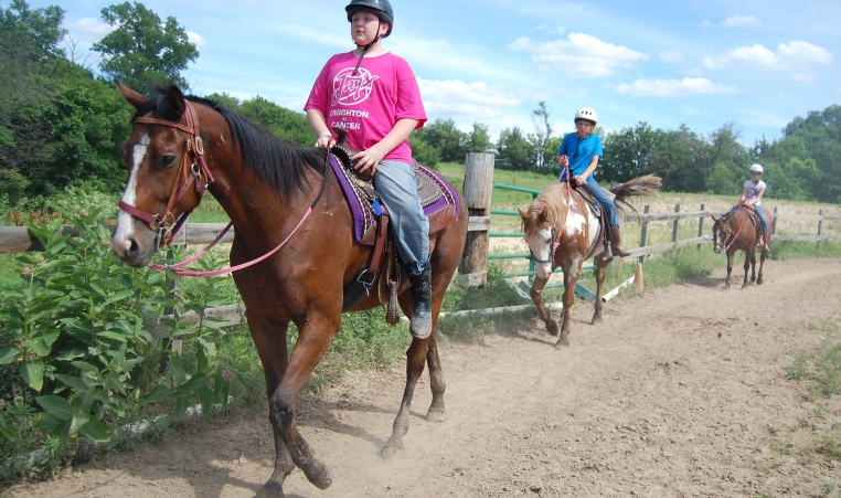 Three campers ride horses along the arena fence line under a blue sky