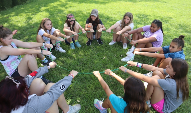 A group of kids sit in a circle leaning back against a loop of rope using it to stand as a team at camp.  