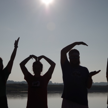 Four people make the letters YMCA with their arms as silhouettes in front of the camp lake