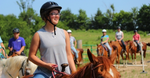 A camper with glasses on horseback smiles while other campers on horseback follow her lead.